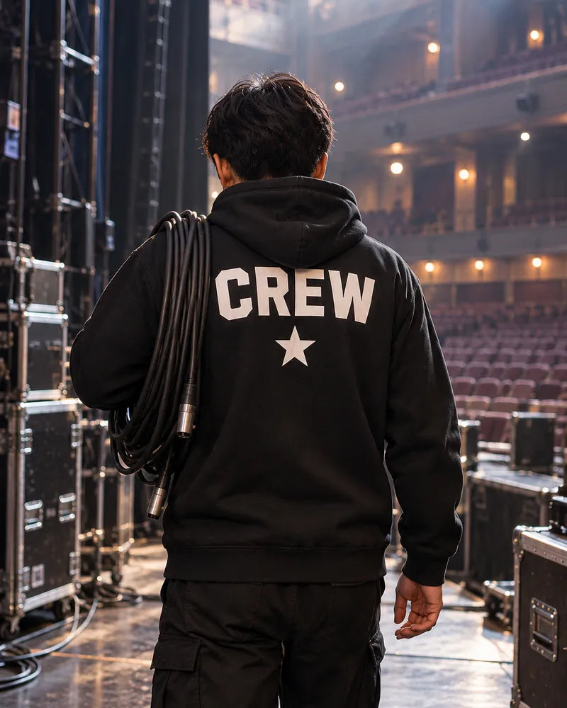 ライブハウスの舞台袖で、背中にCREWと星をプリントした黒いパーカーのスタッフがケーブルを肩にかけている後ろ姿