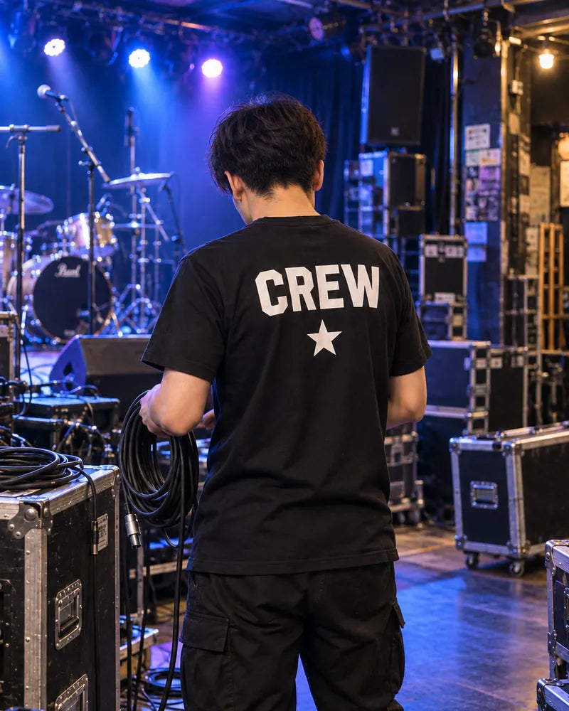 ライブハウスで、背中にCREWと星をプリントした黒Tシャツのスタッフがケーブルを持っている後ろ姿