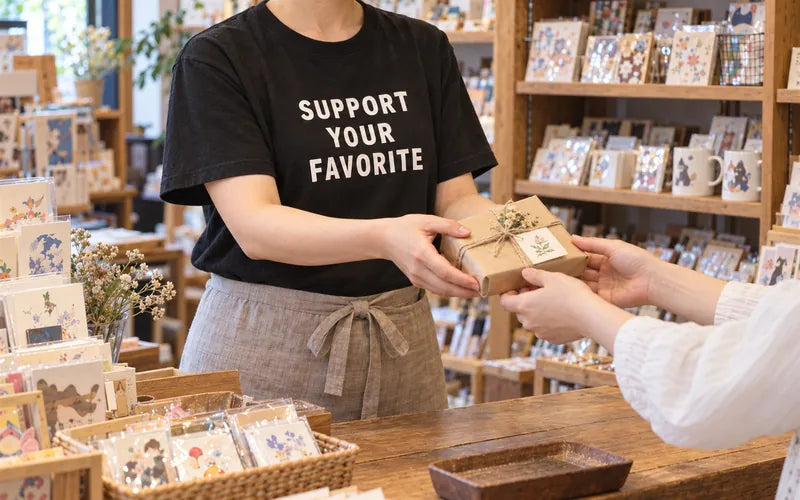 雑貨店で、SUPPORT YOUR FAVORITEの黒Tシャツを着た店員が、お客さんに商品を手渡している